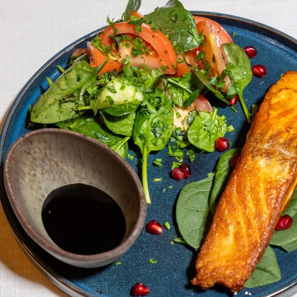 A golden-brown salmon fillet with a fresh spinach and tomato salad, pomegranate seeds, and a dark sauce.